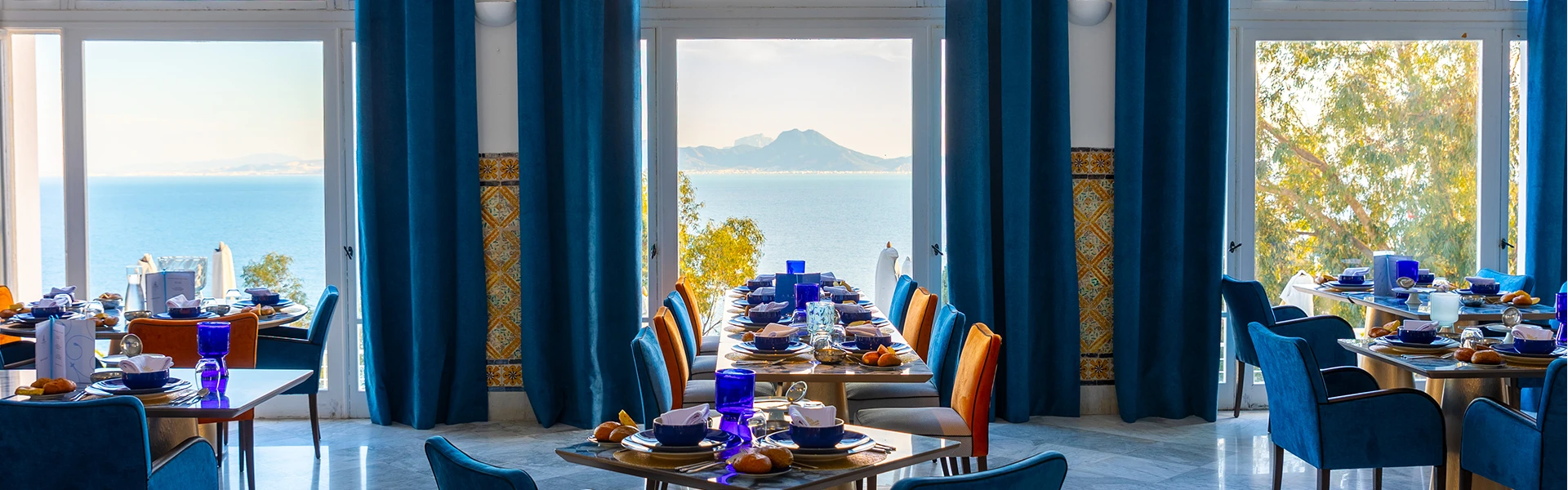 Salle du restaurant aux grands rideaux bleus, tables dressées et baies vitrées ouvertes sur la mer