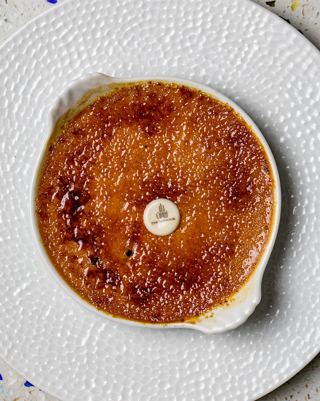 Crème brûlée caramélisée dans un petit plat blanc, vue de dessus, avec le logo Dar Zarrouk