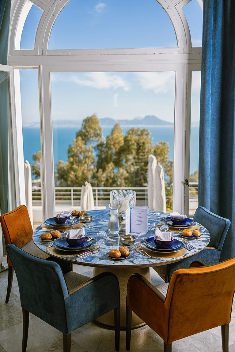 Table ronde dressée devant une grande fenêtre cintrée avec vue sur la baie et les montagnes